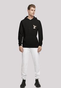 Svart hettegenser med snøring og frontlomme, med et tegneseriehundedesign på brystet; sammen med hvite joggebukser og svarte sko.