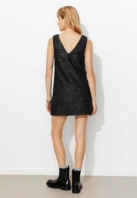 Robe noire sans manches en tissu texturé scintillant, dos en V, longueur au-dessus du genou, associée à des bottines noires et des chaussettes grises.