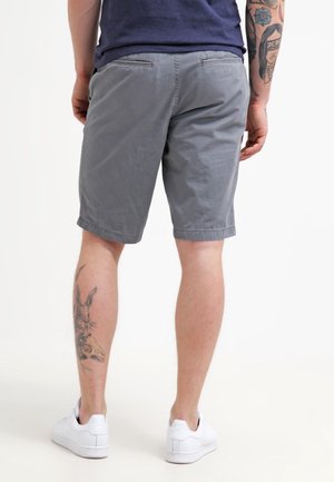 Mand iført grå shorts, hvide sneakers, med tatoveringer på venstre læg og højre underarm, står med ryggen til kameraet mod en hvid baggrund.