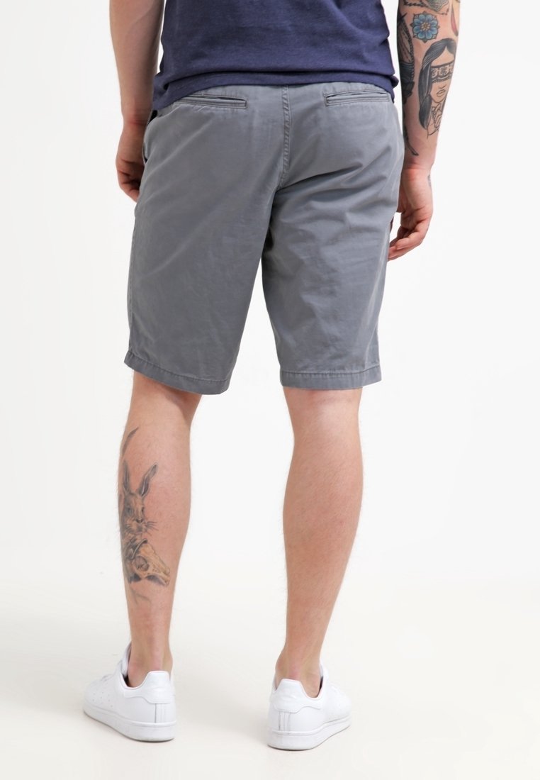 Homme portant un short gris, des tennis blanches, avec des tatouages sur le mollet gauche et l'avant-bras droit, se tenant contre un fond blanc, dos à l'appareil photo.