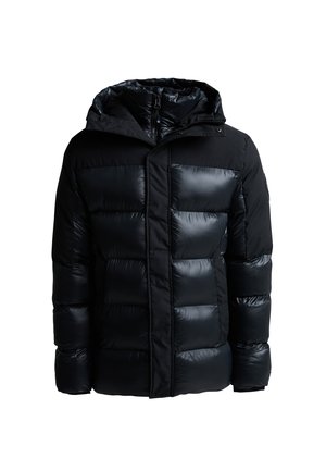 Jonge man die een zwarte gewatteerde jas, zwarte hoodie, zwarte broek en zwarte sneakers draagt, staand tegen een lichtgrijze achtergrond.