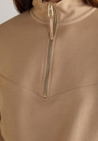 Sudadera beige con cremallera, cuello alto, cremallera dorada y costuras en contraste en el pecho. Textura de tela suave y lisa.