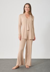 Μπεζ σετ καθιστικού που περιλαμβάνει ένα ανοιχτό μανίκι μακρύ cardigan, μια αμάνικη μπλούζα και καμπάνα παντελόνια φτιαγμένα από απαλό, ελαστικό ύφασμα με ομαλή υφή.