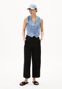 Denim vest met een V-halslijn en vijf metalen knopen, gecombineerd met zwarte wijde broek en zwarte slide-sandalen. Lichtblauwe pet op het hoofd.