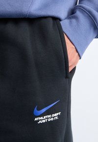 Sorte bomullsbukser i svart med sidelomme, med en blå Nike-logo og hvit tekst som leser "ATHLETIC DEPT JUST DO IT."