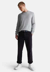 Joven vestido con una camisa de manga larga gris clara, pantalones negros arremangados con un cinturón azul, calcetines blancos y zapatillas blancas, de pie mirando hacia abajo.