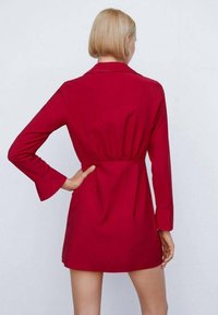 Robe chemise rouge avec manches longues, taille ceinturée et col. Tissu lisse et poignets évasés. Design longueur genou, style minimaliste.