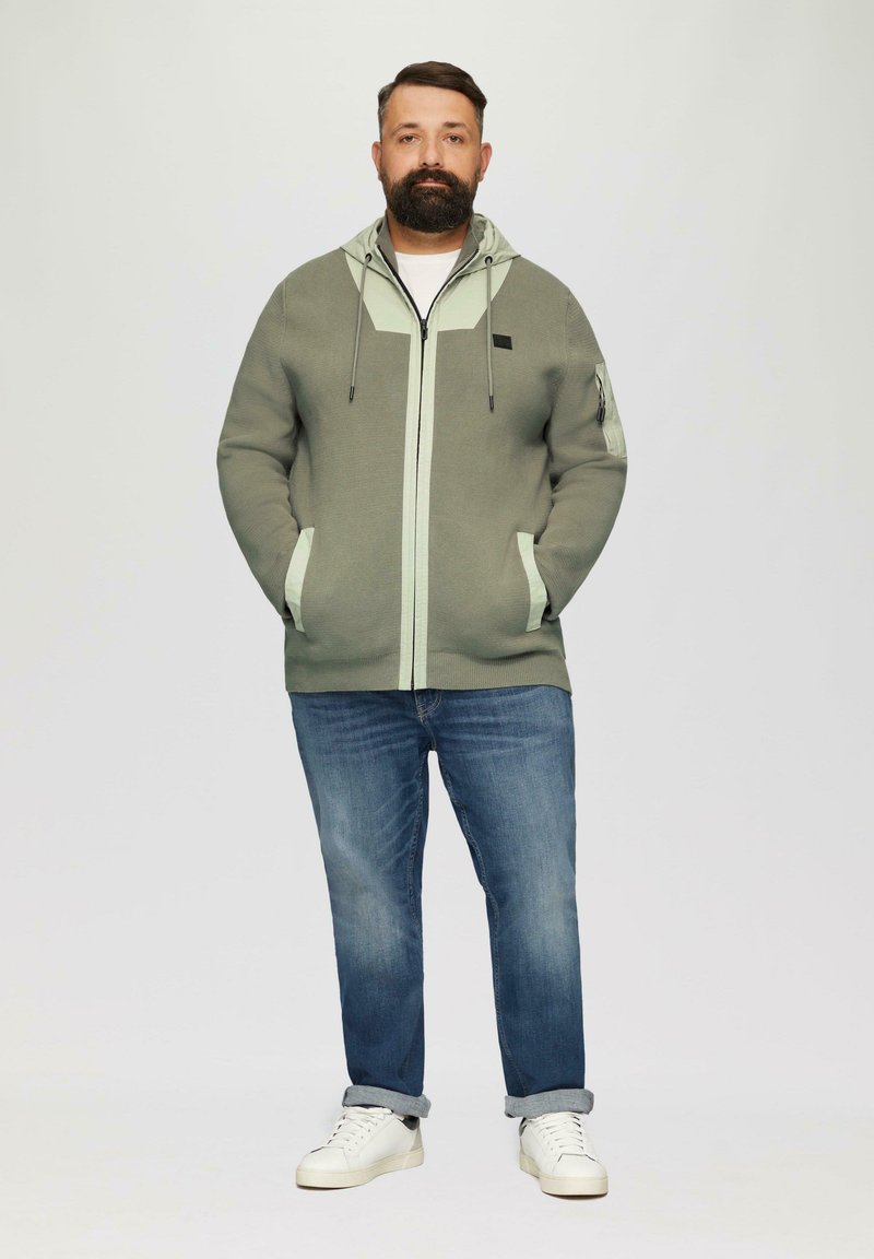 Light green knitted hoodie with contrasting light mint zippers, side pockets, and a hood, paired with blue jeans and white sneakers.