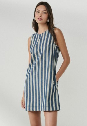 Mladá žena s dlhými tmavými vlasmi, oblečená v modro-bielych vertikálne pruhovaných šatách bez rukávov s vreckami, stojaca pred jednofarebným pozadím.