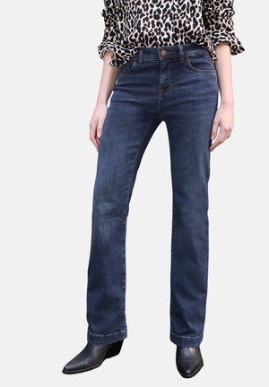 Femme portant un jean droit bleu foncé, des bottes noires à bout pointu et un chemisier à manches longues imprimé léopard avec les manches retroussées.