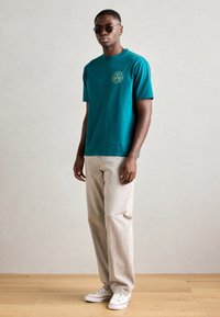 Tyrkis bomulds T-shirt med rund halsudskæring og korte ærmer, der har et rundt grafisk motiv. Paarret med lysegrå denim bukser og hvide sneakers.