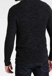 Svart stickad tröja med en strukturerad mönster, rundad halsringning och långa ärmar. Bärs över mörka denimjeans, som visar ribbade muddar och nederkant.