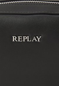 Svart syntetisk materiale med teksturert overflate, med en sølvfarget "REPLAY"-logo som er tydelig vist. Glidelås lukking på toppen.