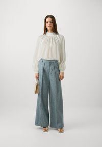 Hvit, mykt teksturert bluse med høy hals; løst-fittende, lyseblå vide bukser; holder en beige veske; flate sandaler.