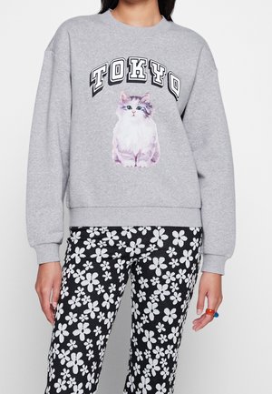Grå sweatshirt med teksten "TOKYO" over en illustration af en hvid og grå kat, båret med sorte bukser med store hvide blomsterprint.