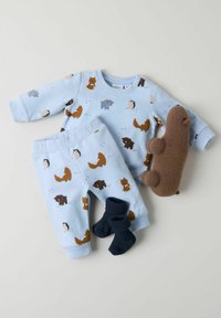 Gaiši zila bērnu pidžamu komplekts ar dzīvnieku rakstiem brūnā, pelēkā un oranžā krāsā, kopā ar tumši ziliem zeķēm un brūnu mīksto rotaļlietu.