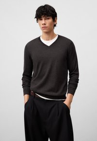 Dunkelgrauer Wollpullover mit V-Ausschnitt, über einem weißen Hemd getragen, kombiniert mit schwarzen, plissierten Hosen. Glatte Textur, enganliegendes Design, lässiger Stil.