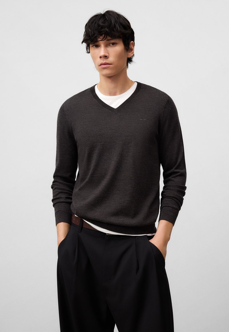 Dunkelgrauer Wollpullover mit V-Ausschnitt, über einem weißen Hemd getragen, kombiniert mit schwarzen, plissierten Hosen. Glatte Textur, enganliegendes Design, lässiger Stil.