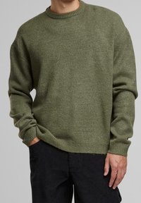 Olivově zelený pletený svetr s kolem krkem, vroubkovanými manžetami a lemem. Texturovaná tkanina, volný střih, nošený s černými kordurovými kalhotami.