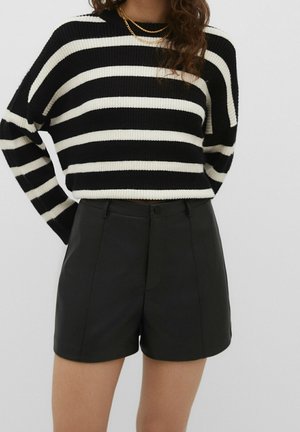 Vrouw draagt een zwart-wit gestreepte trui, zwarte shorts met hoge taille en twee gouden kettingen tegen een effen achtergrond.