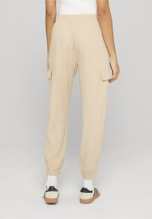 Person wearing beige cargo jogger pants with elastic cuffs, paired with white socks and cream sneakers with black stripes, facing away.