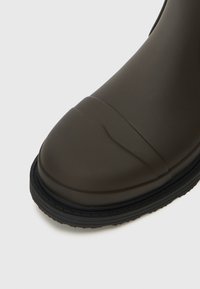 Botte en caoutchouc marron foncé avec une pointe arrondie, semelle antidérapante texturée et détails subtils le long du capuchon. Surface supérieure lisse.