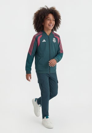 Barnet går og smiler iført en mørkegrøn Real Madrid-jakke med lyserøde striber, matchende cargobukser og hvide sneakers på en hvid baggrund.