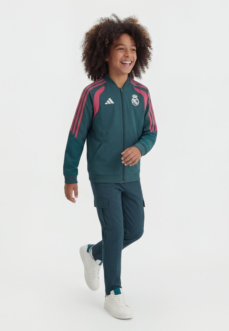Enfant marchant et souriant, portant une veste Real Madrid vert foncé avec des bandes roses, un pantalon cargo assorti et des baskets blanches sur fond blanc.