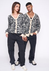 Grau- und cremefarbene gemusterte Cardigans mit schwarzen Knöpfen, getragen über schwarzen Cargo-Hosen und Sneakers. Die Stoffe wirken weich und strukturiert.