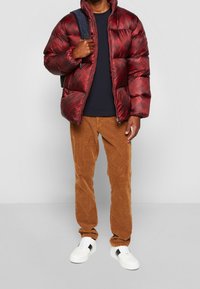 Punainen puffatakkia, jossa on geometrinen kuvio, edessä zip, ja korkea kaulus. Yhdistetty ruskeisiin corduroy-housuihin ja valkoisiin tennareihin, joissa on mustia yksityiskohtia.