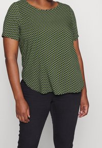 Kvinna i en grönmönstrad kortärmad topp och svarta byxor, stående mot en enfärgad ljus bakgrund, med överkroppen synlig.