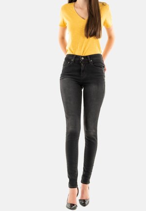 Jeans negros ajustados con cintura alta y bolsillos frontales, combinados con una camiseta amarilla de cuello en V y mangas cortas, llevados con tacones negros.