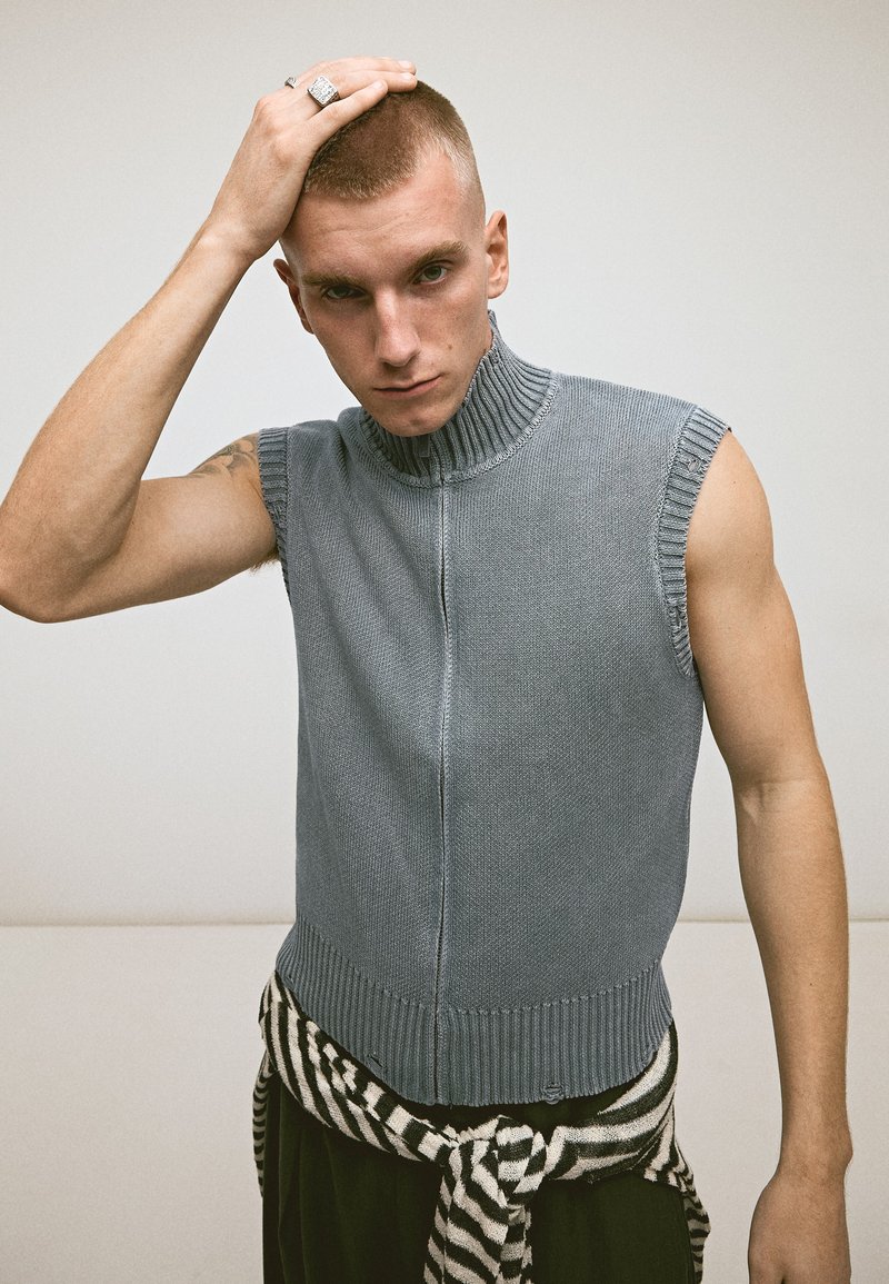Jeune homme aux cheveux courts portant un haut en tricot gris sans manches et un pull rayé noué autour de la taille, main reposant sur la tête.