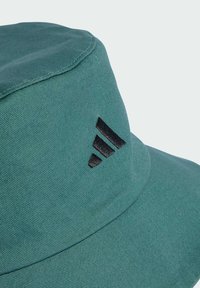 Zelený klobúk typu bucket vyrobený z textúrovanej tkaniny, s čiernym logom Adidas na boku a okrúhlym, širokým okrajom.