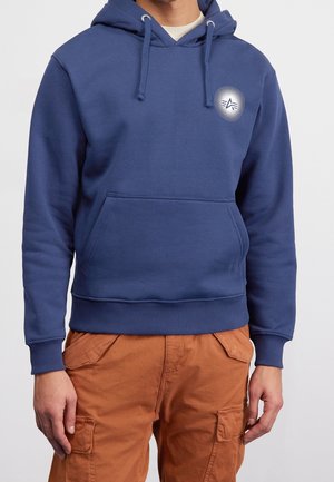 Man wearing blue hooded sweatshirt with front pocket and white circular logo on chest, paired with brown cargo pants.