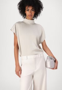 Licht beige top met hoge kraag en korte mouwen, gecombineerd met witte netjes gesneden broek. Houdt een kleine zilveren satijnen pouch vast. Over het geheel een gladde textuur.