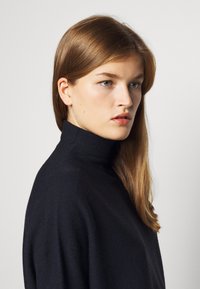 Marineblå turtleneck-genser med en myk tekstur, med høy krage og avslappet passform. Fanget i profil mot en ensfarget bakgrunn.