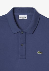 Polo de Lacoste de color navy con cuello clásico, dos botones negros y un logotipo de cocodrilo verde bordado en el pecho. Tejido texturizado.