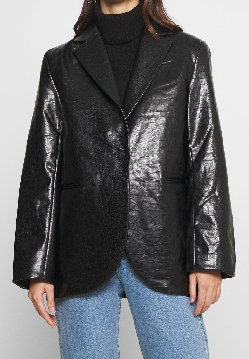 Femme portant un blazer en similicuir noir texturé avec un seul bouton, associé à un col roulé noir et un jean bleu clair.