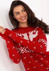 Lächelnde Frau mit langen braunen Haaren, die einen roten Weihnachtspullover mit weißen Rentier- und Schneeflockenmuster trägt und mit zusammengeschlungenen Händen posiert.