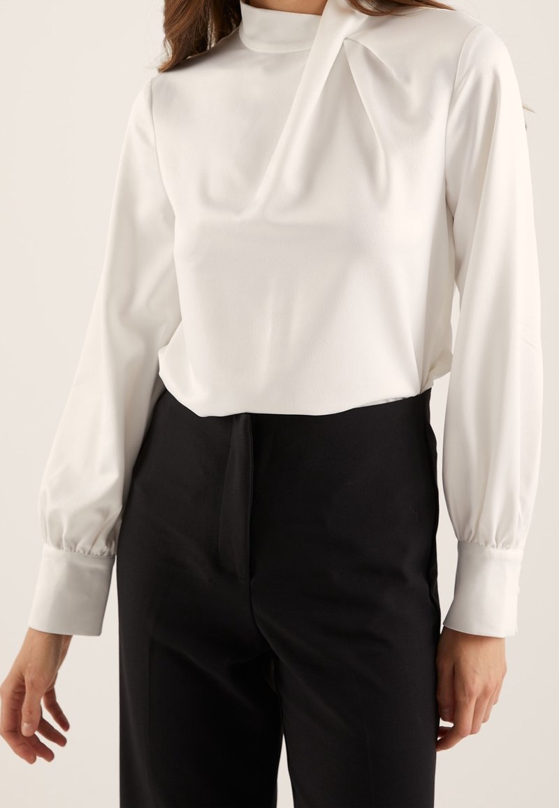 Femme portant un chemisier blanc à manches longues avec poignets retroussés et un pantalon noir taille haute, posant devant un fond uni.