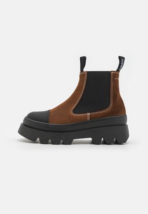 Bota de tornozelo em camurça castanha com ponta de borracha preta e sola grossa e estriada. Painéis elásticos nas laterais e laço na parte de trás. Detalhes costurados por toda a parte.