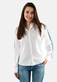 Camisa blanca de botón con mangas largas y acentos de encaje azul. La tela tiene una textura suave y el corte es ajustado y contemporáneo.