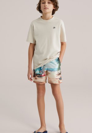 Bomuld T-shirt i creme med en lille broderet logo; farverige badeshorts med strandtema med palmer og både; båret med blå sandaler.