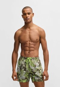 Men's swim trunks featuring a brown base with large green and white tropical leaf patterns, elastic waistband, and drawstring closure.