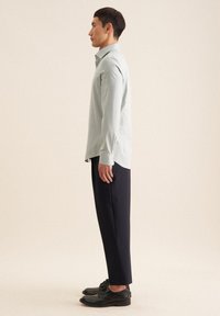 Lichtgroen overhemd met lange mouwen en kraag, gecombineerd met donkerblauwe broek en zwarte nette schoenen, vanuit de zijkant getoond.