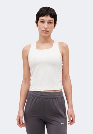 Vrouw met kort donker haar, draagt een wit mouwloos shirt en grijze Nike sportbroek tegen een effen lichte achtergrond.