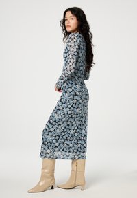 Blauwe bloemen jurk met lange mouwen en een getailleerd silhouet, gemaakt van doorschijnende stof met een zwarte onderlaag; gecombineerd met beige gepatineerde kniehoogte laarzen.