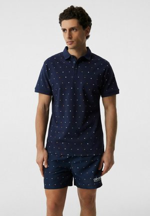 Mężczyzna w granatowej koszulce polo i pasujących szortach z małymi kolorowymi geometrycznymi wzorami, stojący na jednolitym tle.