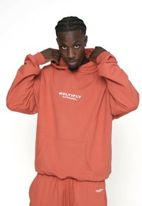 Bărbat cu coada de pește își ajustează gluga unui hanorac coral "Multiply Apparel" și pantalonilor asortați pe un fundal alb.
